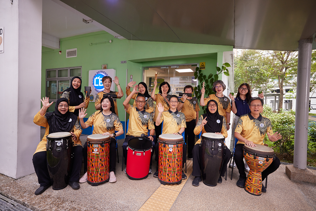 Group of people with drums and percussion instruments wearing Lions Befrienders t-shirts, waving.